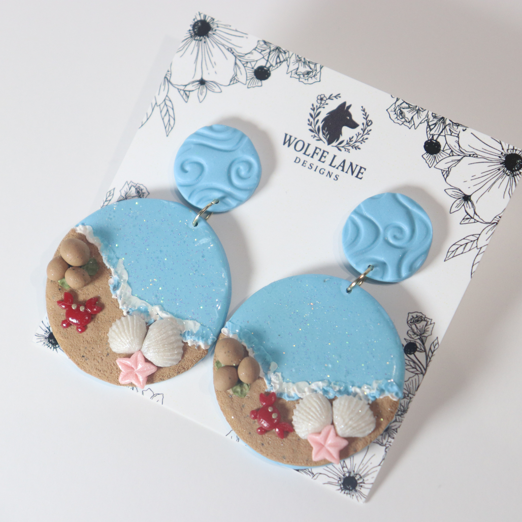 Pair of blue earrings with beach-themed designs on a Wolfe Lane Designs card.